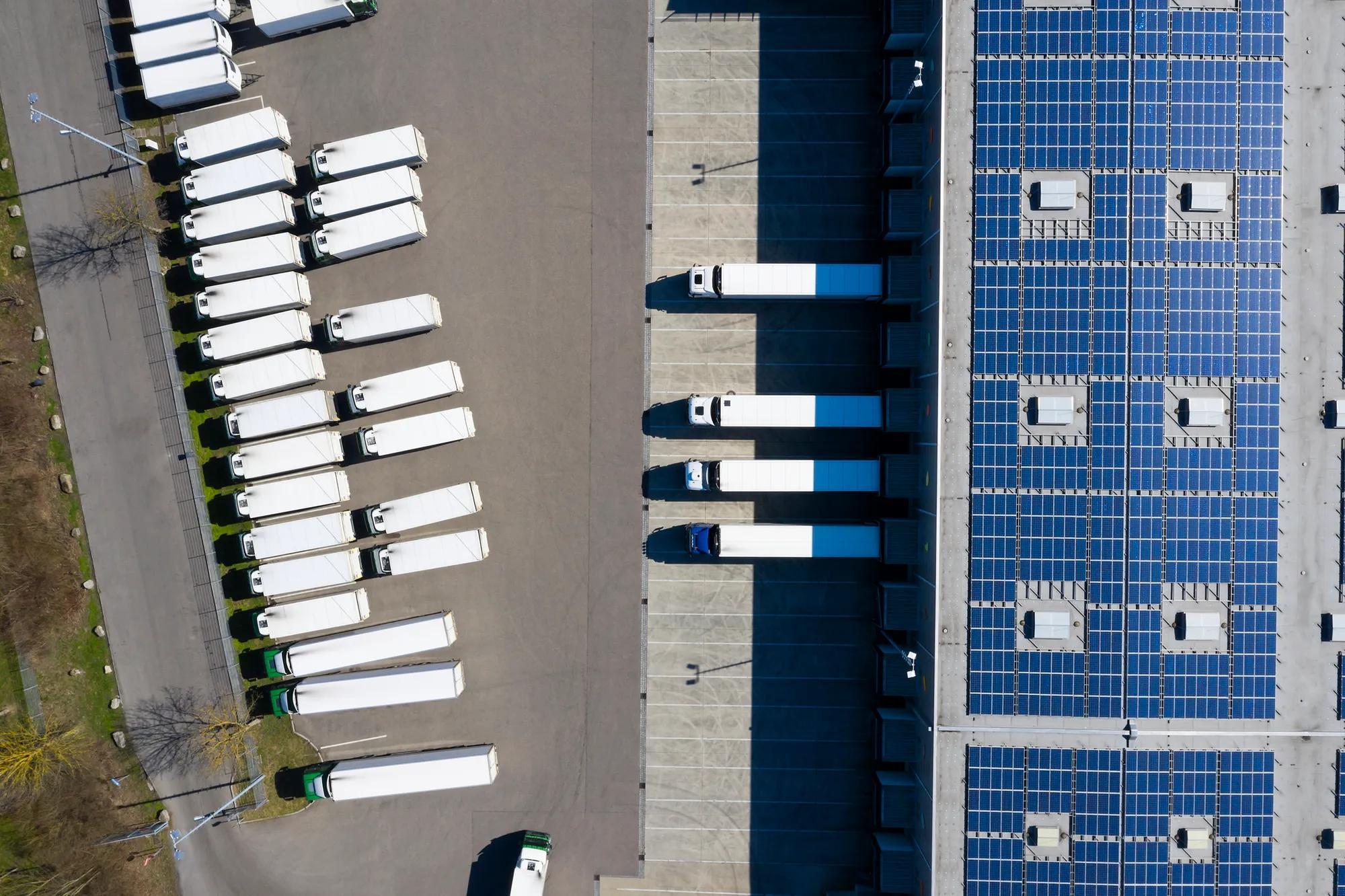 Vue aérienne de camions en cours de déchargement et d'un grand entrepôt avec panneaux solaires sur le toit. 